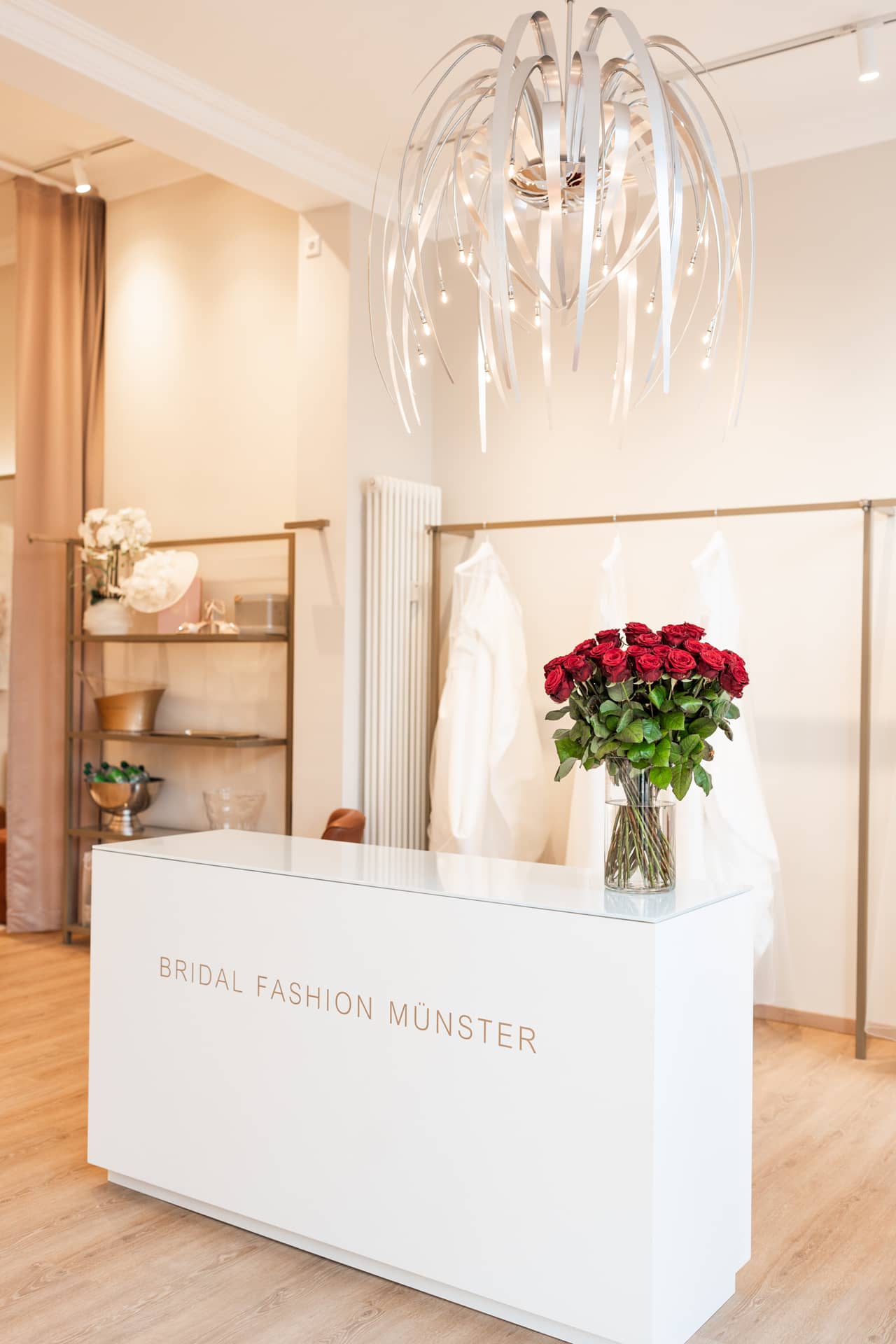 anabela-privee-laden-innen-theke Rezeptionstresen von Bridal Fashion Münster mit einem Strauß roter Rosen und moderner, spiralförmiger Deckenbeleuchtung.