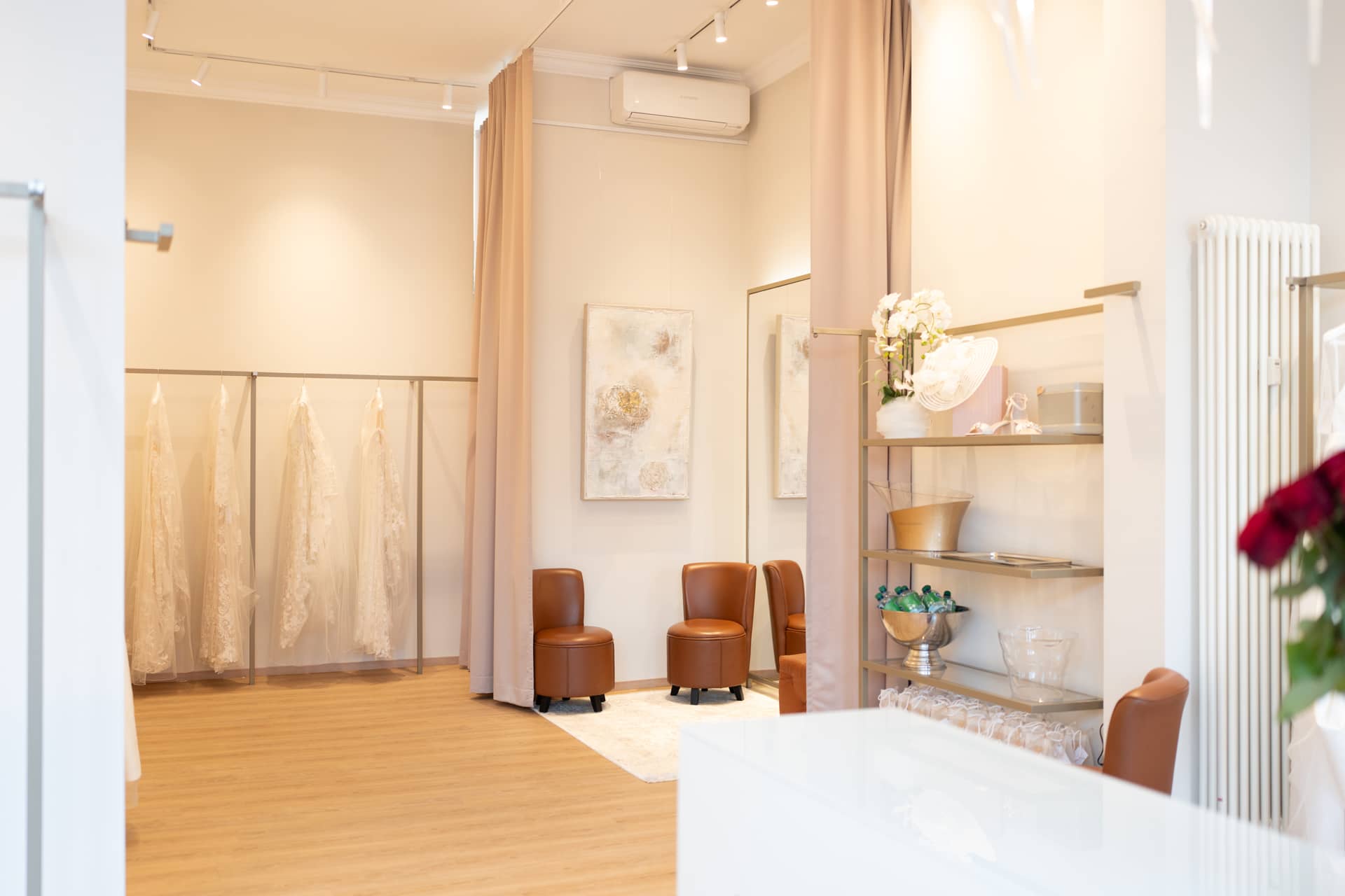 anabela-privee-laden-innen Eleganter Brautmodenladen mit Hochzeitskleidern auf Kleiderständern, gemütlichen Sesseln und einer dekorativen Vase mit weißen Blumen auf einem Regal.